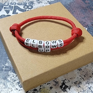 May include: Red paracord bracelet with white beads spelling "ELBOWS UP".