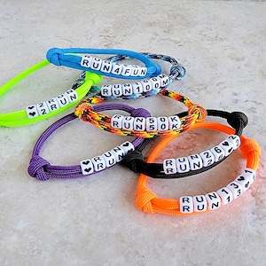 May include: A collection of five adjustable paracord bracelets in neon green, blue, multicolored, purple, and orange. Each bracelet features white square beads with black text including RUN 4 FUN, RUN 100M, RUN 50K, RUN, and RUN 26.2 or RUN 13.1, accented with small black heart symbols.
