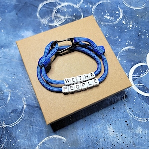 May include: A blue rope bracelet with the words "WE THE PEOPLE" spelled out in white letter beads. The bracelet is on a brown cardboard box, set against a blue background.