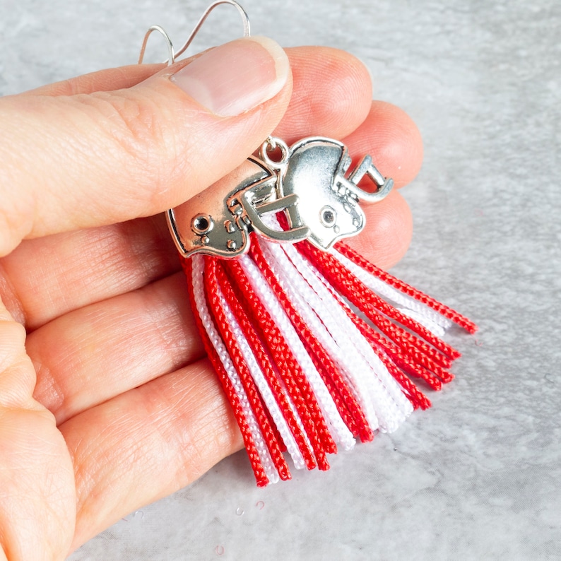 close up of a hand holding red and white tassel earrings with silver football helmet charms