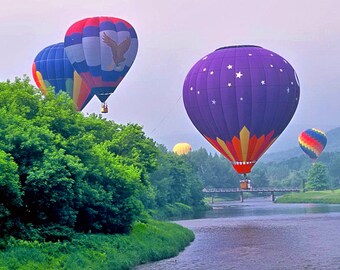 May include: Four hot air balloons in the sky, two are red, white, and blue, one is purple with white stars, and one is rainbow colored. The balloons are flying over a river and green trees.