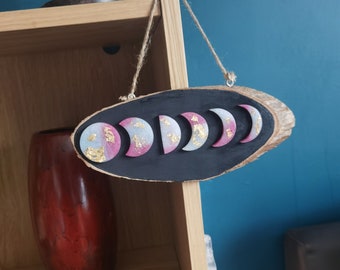 Pode incluir: Um pendurado de parede de madeira com fases da lua, fundo preto e luas pintadas de rosa e branco. As luas têm detalhes dourados e estão dispostas em forma de crescente.
