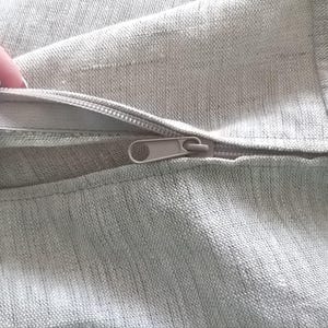 May include: A close-up of a zipper on a light gray fabric cushion cover. The zipper is open and the fabric is soft and textured.