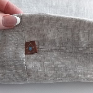 Puede incluir: Un primer plano de una tela de lino gris claro con un parche de cuero marrón cosido. El parche tiene una casa blanca con una flor azul dentro.