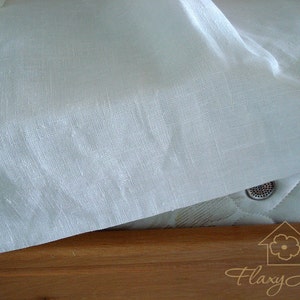 May include: A white linen fabric sheet draped over a wooden surface. The fabric has a subtle texture and a slightly crinkled appearance. The surface is a light brown color and has a smooth finish.