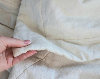 Könnte beinhalten: Nahaufnahme eines cremefarbenen Stoffes mit sichtbarer Textur und Nähten. Der Stoff scheint ein natürliches Material zu sein, möglicherweise Leinen oder Baumwolle, mit einem leicht zerknitterten Aussehen. Das Bild hebt die subtilen Farb- und Texturvariationen des Stoffes hervor.