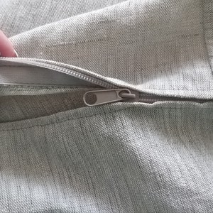Puede incluir: Primer plano de una cremallera en una funda de coj&iacute;n de tela gris claro. La cremallera est&aacute; abierta y la tela es suave y texturizada.