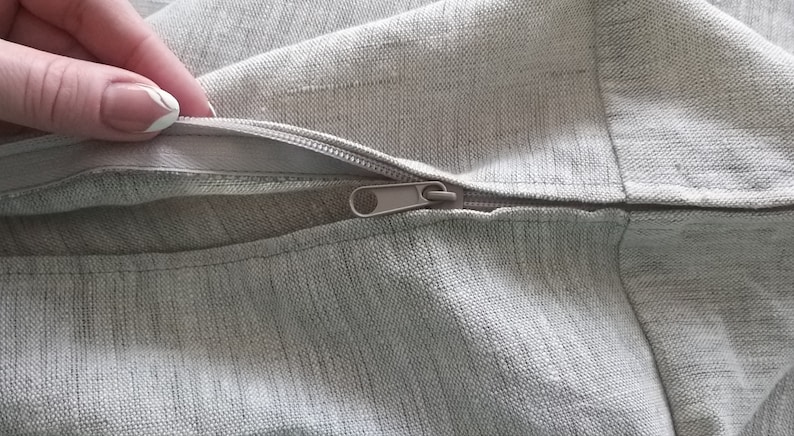 Puede incluir: Primer plano de una cremallera en una funda de coj&iacute;n de tela gris claro. La cremallera est&aacute; abierta y la tela es suave y texturizada.