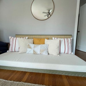 Op de afbeelding: Een witte futon met een groene hoofdbord en een witte matras. De futon is versierd met kussens in verschillende kleuren en patronen, waaronder rood-wit gestreept, beige, geel en wit met een geometrisch patroon.