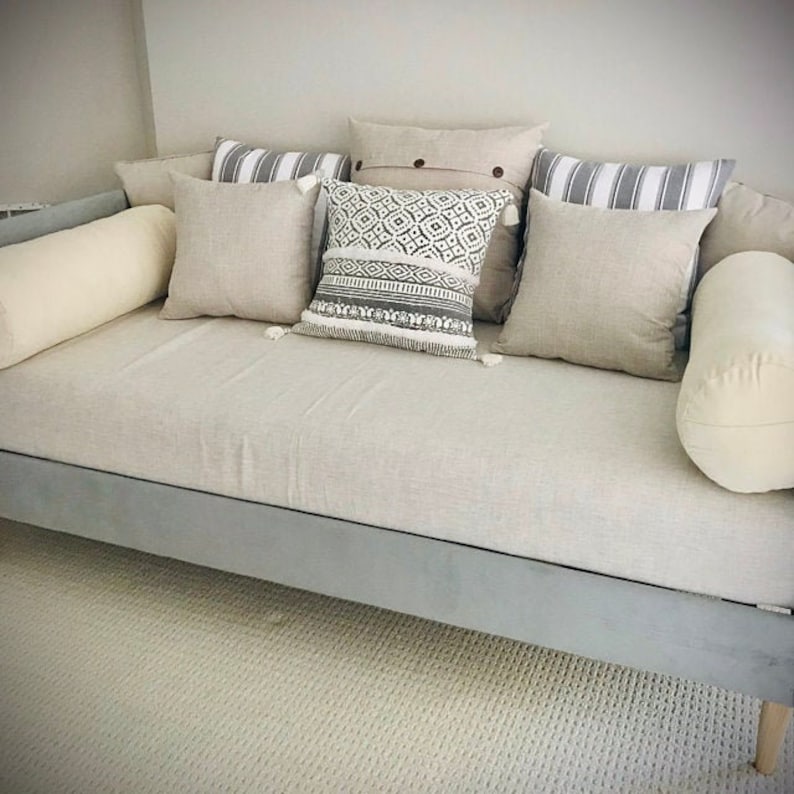 Op de afbeelding: Een lichtgrijze dagbed met een witte linnen bekleding en verschillende kussens. De kussens zijn in verschillende tinten beige en wit met geometrische patronen. De dagbed heeft een grijs houten frame en poten.