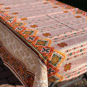 Puede incluir: Un mantel rectangular con fondo beige y un patrón repetitivo de rayas verticales. El borde presenta un diseño geométrico en naranja, verde y marrón. El mantel está hecho de tela tejida.