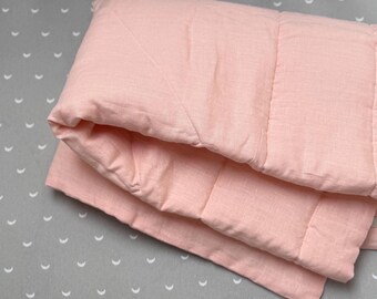 May include: A folded, light pink, quilted blanket. The blanket is made of a soft, textured fabric and has a stitched pattern. The background is a gray fabric with a white crescent moon pattern.