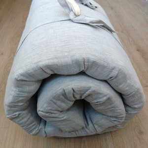 May include: A grey linen futon mattress rolled up and tied with a white ribbon. The mattress is made of a soft, comfortable material and is ideal for a spare room or a small flat.