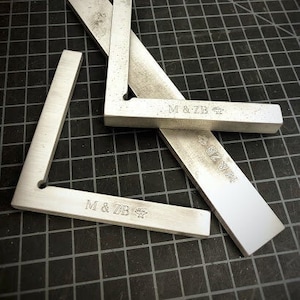 May include: Two silver metal right angle rulers with the text "M & ZB" engraved on each. The rulers are on a black and white grid surface.