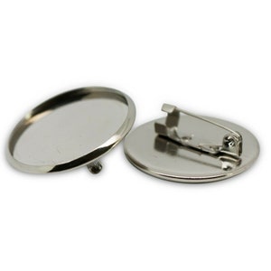 May include: Two silver-coloured, oval-shaped pin backs. One is face up, showing the flat surface for attaching a design. The other is face down, revealing the pin and clasp mechanism. Suitable for custom badges or pins.