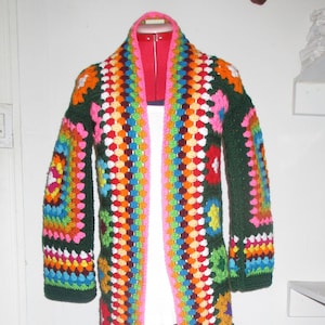 May include: A long, crocheted cardigan with a colorful geometric pattern and green fringe. The cardigan is open in the front and has a white lining.