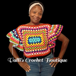 Könnte beinhalten: Ein mehrfarbiges Häkeloberteil mit quadratischem Granny-Stitch-Muster in leuchtenden Farben wie Orange, Gelb, Pink, Blau und Violett. Das Kleidungsstück hat weite, gerüschte Ärmel und einen U-Boot-Ausschnitt. Der Text Valli's Crochet Boutique ist sichtbar.