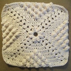 May include: A white crocheted square with a raised, textured pattern. The center of the square features a larger, more intricate flower design.