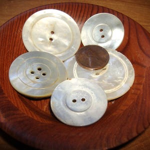 May include: A collection of five round, off-white mother-of-pearl buttons with varying hole counts, including two-hole and four-hole designs. A copper-colored United States penny is placed on top of the buttons for size comparison.