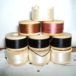 May include: A pyramid stack of eight wooden spools of thread in various colors including white, black, copper, and mauve. Each spool features a light-colored wooden core with thread wound around the center, and some spools have visible text.