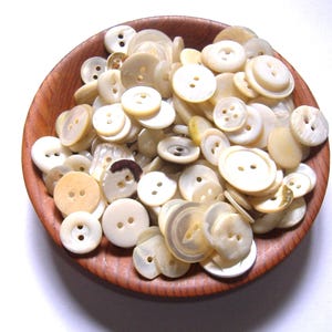 May include: A collection of assorted round off-white and cream-colored buttons with two or four holes, contained within a small, circular, polished wooden bowl. The buttons vary in size, thickness, and texture, displaying smooth and slightly iridescent surfaces.