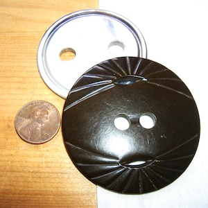 May include: A dark brown circular button with a sunburst-style carved pattern and two central holes, positioned next to a copper-colored United States penny for scale. A second, plain white circular button with two holes is partially visible underneath.