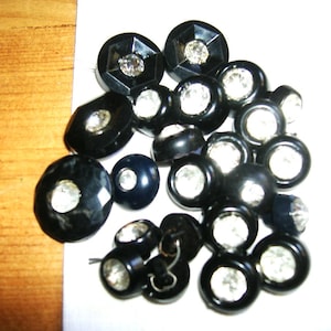 May include: A collection of round black buttons, each featuring a clear, faceted crystal or rhinestone set in the center. The buttons vary slightly in size and shape, with some having smooth edges and others displaying a geometric, multi-faceted design.