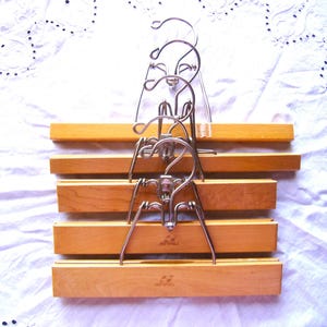 May include: A set of five light-colored wooden pant hangers stacked vertically. Each hanger features a smooth, rectangular wooden bar with a silver-toned metal clamping mechanism and a swivel hook at the top for hanging garments securely.