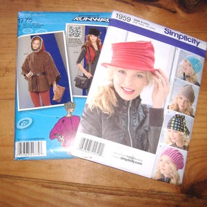 May include: Two Simplicity sewing pattern envelopes. The blue envelope, number 1775, features a brown hooded poncho. The white envelope, number 1959, displays various hat styles including a red hat, a blue beanie, a brown beanie, and a pink knit cap.