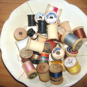 May include: A collection of vintage wooden sewing thread spools in various colors including pink, blue, yellow, black, and brown, contained within a white scalloped ceramic bowl. The spools feature paper labels with brand names.
