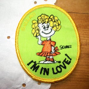 May include: Oval-shaped patch with a cartoon girl in an orange dress and yellow hair, with the text "I'M IN LOVE!" in black. The patch has a green background and a gold border, with the artist's signature.