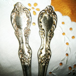 May include: Two antique silver-colored serving spoons with ornate floral and scrollwork detailing. The spoons have a tarnished appearance, suggesting age, and are set against a white, embroidered background.