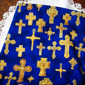 May include: A blue fabric with a pattern of gold-colored crosses. The crosses vary in design and size, with ornate details. A white lace trim borders the fabric, which is displayed on a wooden surface.
