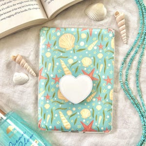 May include: A clear phone case with a seashell and starfish pattern. A white heart-shaped phone grip is attached to the back of the case. The case is lying on a white surface with a turquoise necklace and seashells.