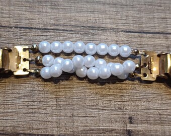 May include: A gold-coloured metal clasp featuring two rows of white faux pearls. The clasp has two rounded end pieces and a central connecting mechanism. The pearls are strung together, creating a decorative accent.