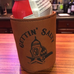 May include: Brown leather can cooler with the text "GETTIN' SALTY" and a black and white illustration of a firefighter's helmet with a moustache.