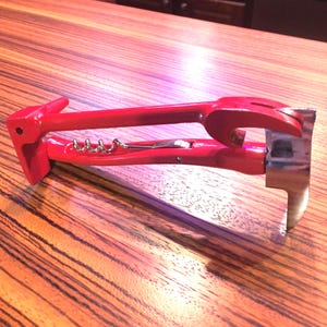May include: A bright red multi-tool shaped like a hammer, featuring a metal corkscrew integrated into the handle, a wrench-style opening on the side, and a silver-coloured metal claw head at the end of the tool, resting on a wood grain surface.