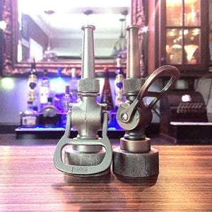 May include: Two metallic bottle openers shaped like vintage fire hose nozzles. The left opener has a silver finish with the word OPEN embossed on the base. The right opener features a darker, bronze-toned finish with a prominent side lever handle.