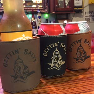 May include: Three brown leather can coolers with the text "Gettin' Salty" and a firefighter helmet graphic. The coolers are on a wooden table with a bottle of beer and two cans of soda in the background.
