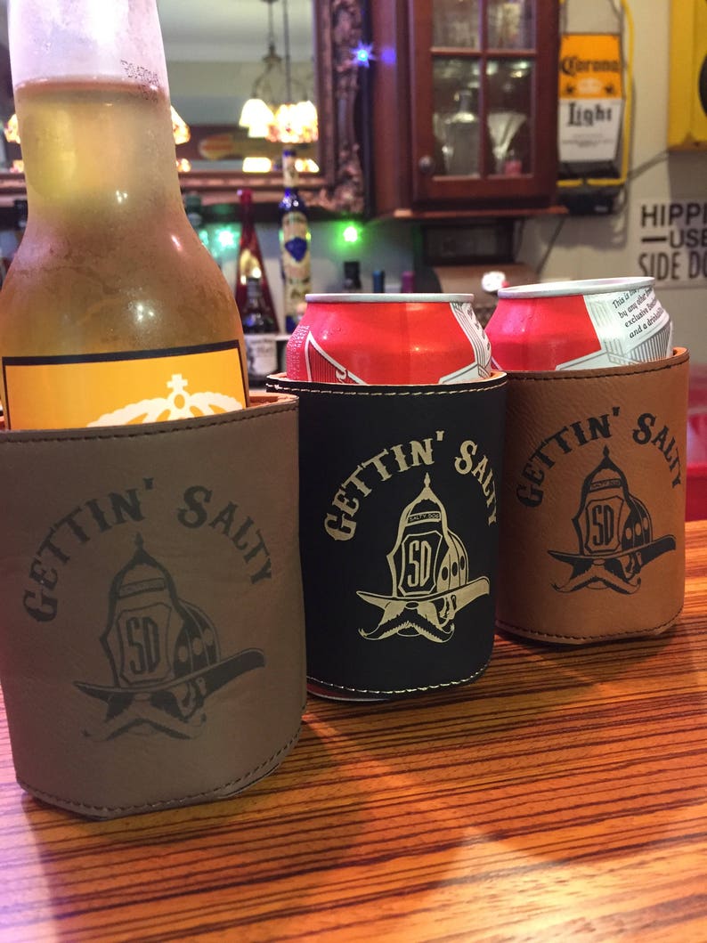 May include: Three brown leather can coolers with the text "Gettin' Salty" and a firefighter helmet graphic. The coolers are on a wooden table with a bottle of beer and two cans of soda in the background.
