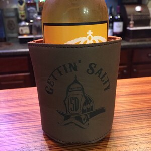 May include: A brown leather beverage holder with the text "Gettin' Salty" and a firefighter helmet graphic. The holder is holding a clear glass bottle with a yellow label.