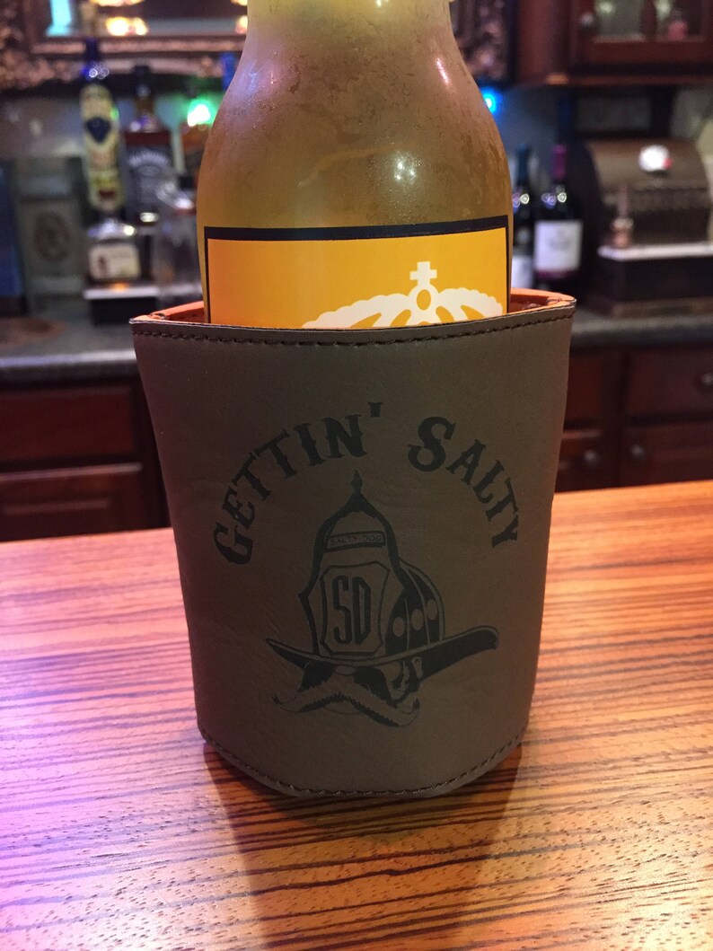 May include: A brown leather beverage holder with the text "Gettin' Salty" and a firefighter helmet graphic. The holder is holding a clear glass bottle with a yellow label.