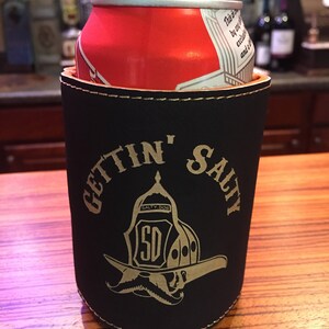 May include: Black neoprene can cooler with a gold stitched border and the text "Gettin' Salty" in gold lettering. A gold firefighter helmet with a moustache and the text "Salty Dog" is in the centre of the cooler.