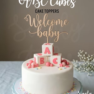 May include: A white cake topped with three white cubes featuring pink letters A, B, and C, surrounded by small pink spheres and star-shaped decorations. A wooden cake topper with the text Welcome baby is positioned above the cubes.