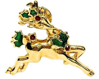 May include: A gold-toned metal brooch shaped like a leaping reindeer. The reindeer features green enamel holly leaves on its antlers and back, accented with small, round, red stones embedded within the holly clusters and on its body.