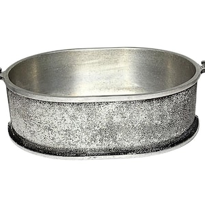 Puede incluir: Un cuenco ovalado de metal con un acabado exterior texturizado y moteado en gris plateado y una superficie interior lisa y pulida. El cuenco presenta dos pequeñas asas decorativas en los lados opuestos, que se extienden desde el borde.