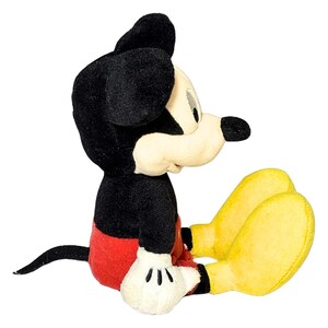 May include: A plush Mickey Mouse toy in a seated position. The toy features black ears, a black body, red shorts, yellow feet, and white gloves. The toy is a classic Disney character.