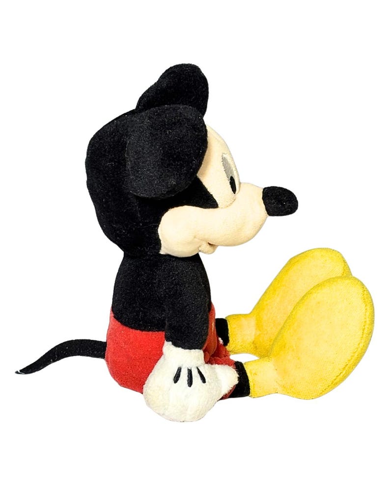 May include: A plush Mickey Mouse toy in a seated position. The toy features black ears, a black body, red shorts, yellow feet, and white gloves. The toy is a classic Disney character.