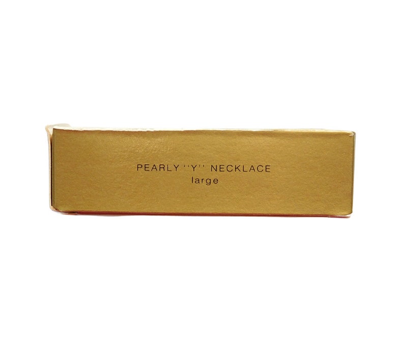 Peut inclure: Une bo&icirc;te dor&eacute;e avec le texte "PEARLY "Y" NECKLACE large" imprim&eacute; dessus.