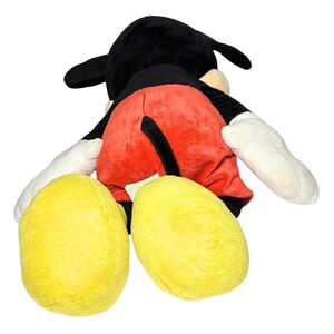 May include: A plush Mickey Mouse toy, viewed from the back. The toy features a black head with large ears, red shorts, yellow feet, and white gloves. The toy is soft and cuddly, perfect for children.